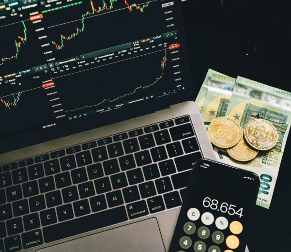 bitcoins and paper money beside a cellphone and laptop with graphs on screen